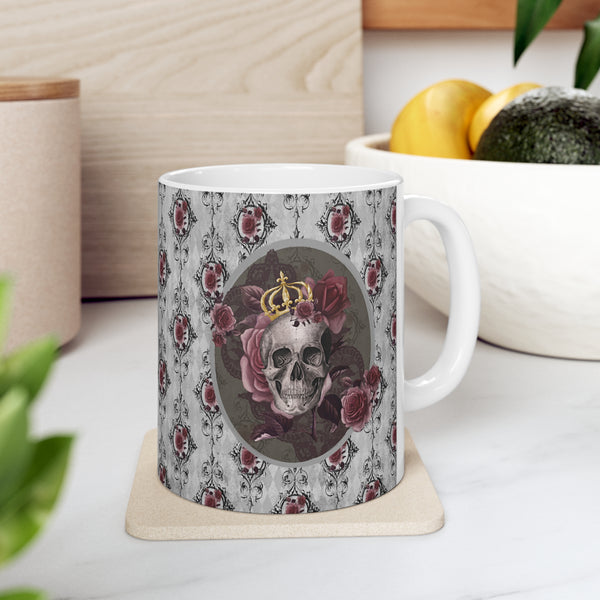 Ceramic mug with a skull wearing a gold crown accented with burgundy roses inside an oval with a light gray border, on a background of gray with black scrolls and burgundy roses with white handle.