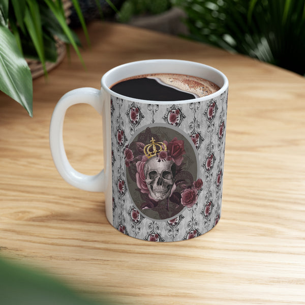 Ceramic mug with a skull wearing a gold crown accented with burgundy roses inside an oval with a light gray border, on a background of gray with black scrolls and burgundy roses with white handle on wood table top.