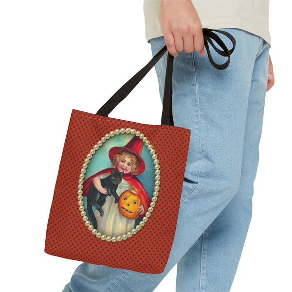 A person in a blue jeans holds a Halloween-themed tote bag with a vintage witch design, featuring a girl with a black cat and a jack-o-lantern, framed by pearls on a polka-dot orange background.