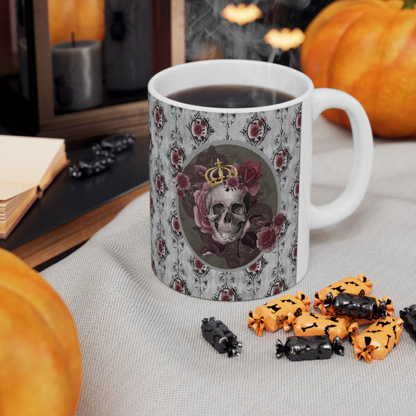 Ceramic mug with a skull wearing a gold crown accented with burgundy roses inside an oval with a light gray border, on a background of gray with black scrolls and burgundy roses with white handle on a table with pumpkin behind and Halloween candy in front of mug.