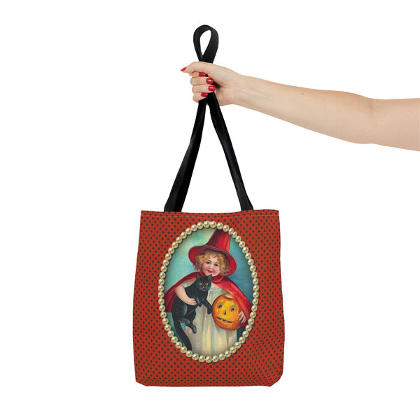 A hand holding a Halloween-themed tote bag with a vintage design of a girl dressed as a witch, holding a black cat and a jack-o-lantern, framed by pearls on a polka-dot orange background.