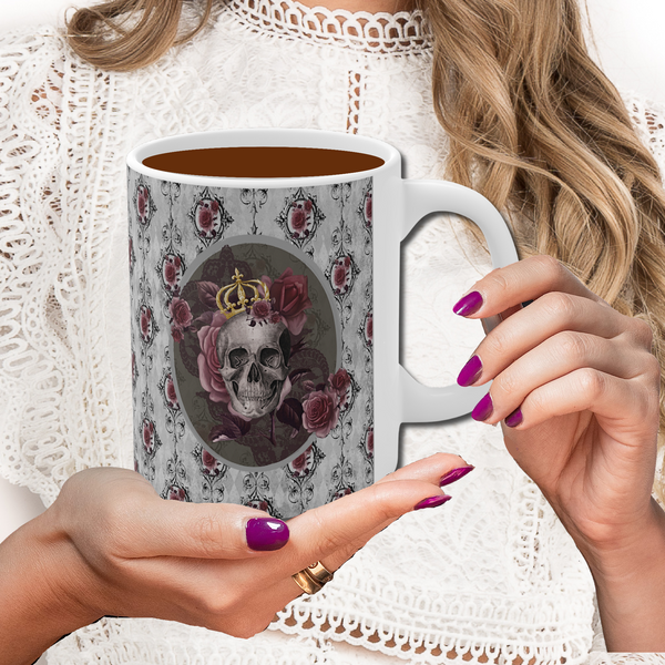 Hands with dark rose nail polish holding a Ceramic mug with a skull wearing a gold crown accented with burgundy roses inside an oval with a light gray border, on a background of gray with black scrolls and burgundy roses with white handle.