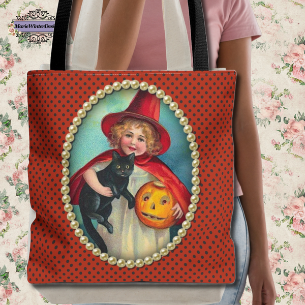 Person holding a Halloween-themed tote bag with a vintage design of a girl dressed as a witch, holding a black cat and jack-o-lantern, framed by pearls on a polka-dot orange background. Floral wallpaper is in the background