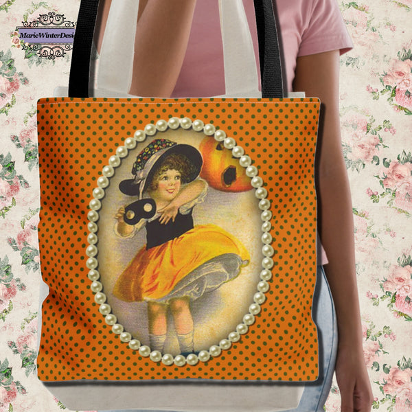 Person holding a tote bag featuring a vintage-inspired Halloween design with a young girl in a witch costume, framed with pearls on an orange polka dot background. Floral wallpaper in the background adds a decorative touch.