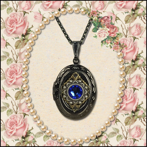 An ornate oval locket pendant necklace with a dark metal frame, a central blue gemstone, and gold-toned filigree details, displayed on a black chain against a vintage floral rose background with a pearl border.