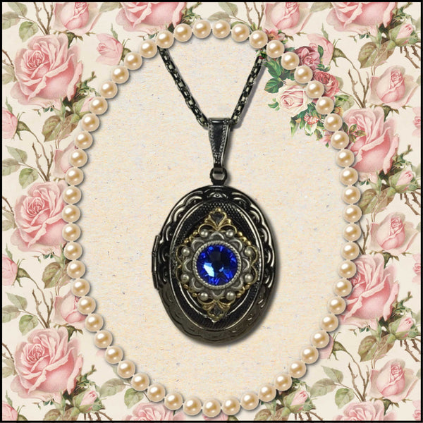 An oval locket pendant necklace with a dark metal frame, ornate filigree detailing, and a central blue gemstone, displayed on a cream background with a pearl strand and pink rose floral border.