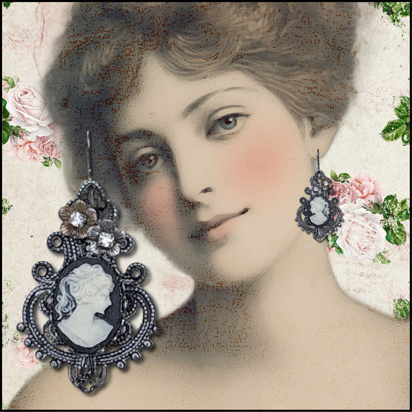 A vintage-style earring displayed over a sepia-toned portrait of a woman with pink cheeks and floral accents around the edges. The earring features an ornate filigree frame with a cameo-style oval inset in white and dark tones, plus small floral details and rhinestone-like stones.
