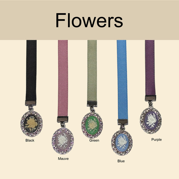 Five oval flower-shaped jewelry pendants with ribbon straps are shown on a beige background under the heading 'Flowers.' The ribbons are labeled Black, Mauve, Green, Blue, and Purple, and the pendant centers depict floral motifs in contrasting colors.