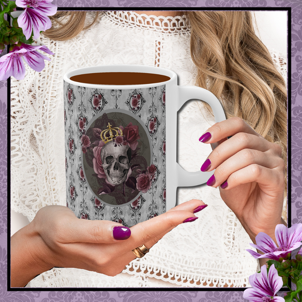 Hands with dark rose nail polish holding a Ceramic mug with a skull wearing a gold crown accented with burgundy roses inside an oval with a light gray border, on a background of gray with black scrolls and burgundy roses with white handle. There is a border of purple with purple flowers in top left and lower right corners.