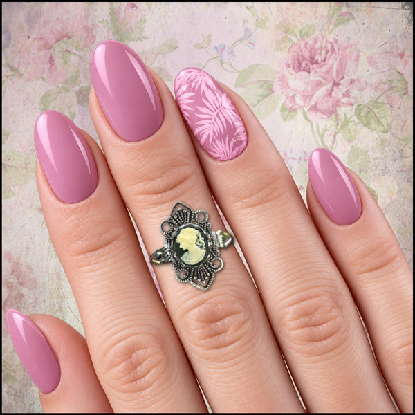 A close-up of a hand with long, almond-shaped pink nails, featuring a pink floral pattern on the ring finger. The hand is wearing an ornate silver-toned ring with a yellow-green cameo-style face inlay, set against a vintage floral background.