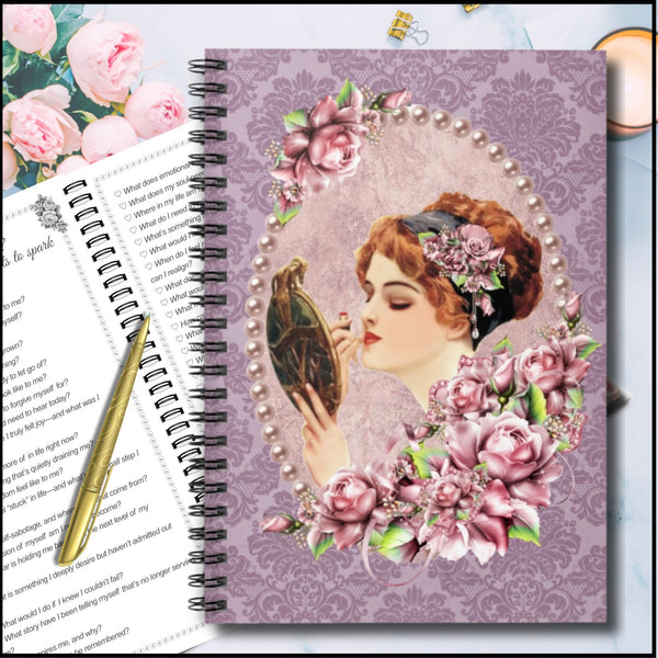 A spiral-bound notebook or journal cover shown in a styled flat-lay. The cover features a vintage illustration of a woman with red hair looking into a mirror, surrounded by pink roses and pearls on a lavender damask background.