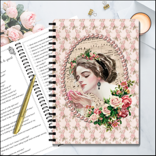 A spiral-bound notebook with a vintage-style floral illustration on the cover, displayed on a light surface with an open book, a gold pen, and small paper clips around it.