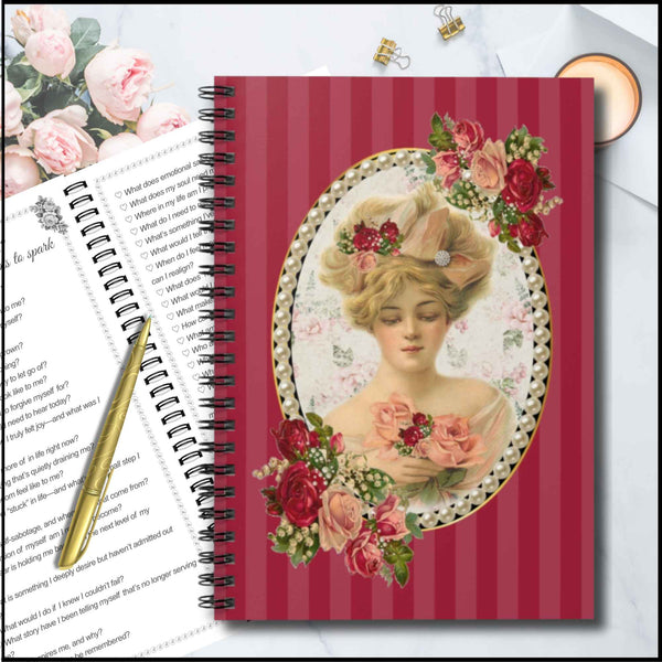 A spiral-bound notebook with a vintage-style floral cover design, shown on a light gray surface with a pen, an open book, and flowers as props.