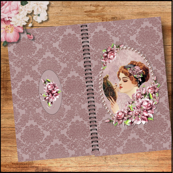 A vintage-style spiral-bound notebook or journal displayed open on a wooden surface. The cover features a dusty mauve damask pattern, with decorative floral and portrait embellishments.