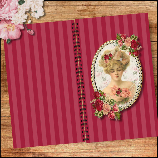A vintage-style spiral notebook with a deep pink and red striped cover, featuring an oval illustration of a woman surrounded by red and pink flowers. The notebook is displayed on a wooden surface with decorative pink flowers in the top-left corner.