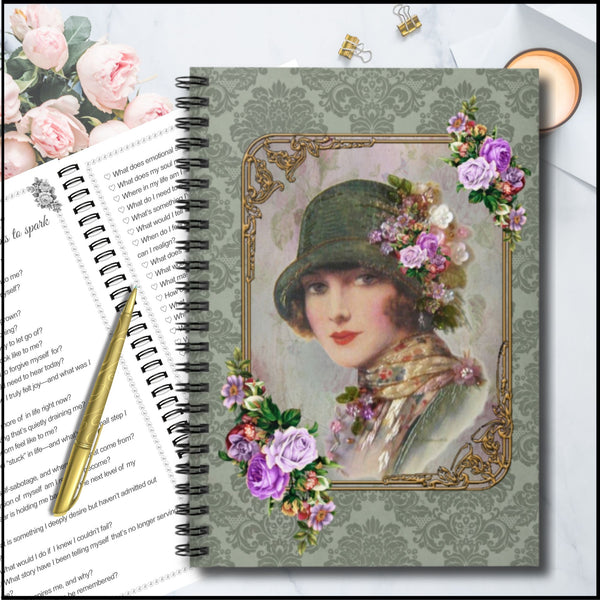 A spiral-bound notebook with a vintage-style cover illustration of a woman in a cloche hat surrounded by purple and pink flowers, shown on a styled desk with an open book, pen, clips, and a candle.