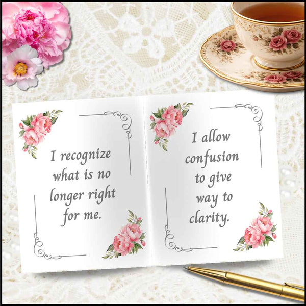 An open white card with pink floral corner illustrations and printed quotes, styled on a lace surface with a teacup, flowers, and a gold pen nearby.