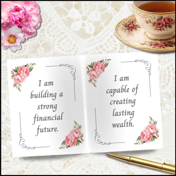 An open white card or notebook spread with pink floral corner illustrations and the printed quote, "I am building a strong financial future." on the left page and "I am capable of creating lasting wealth.