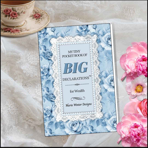 A pocket book titled 'My Tiny Pocket Book of Big Declarations for Wealth' by Marie Winter Designs, featuring a blue floral cover with a white lace border, styled on white lace fabric with pink flowers and a teacup in the background.