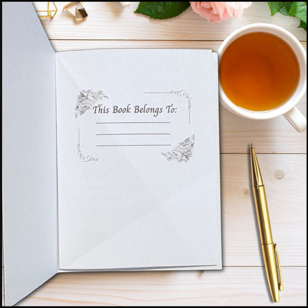 An open white book or folder on a light wood surface with the printed heading 'This Book Belongs To:' and blank lines for writing. A white mug of tea, a gold pen, and small decorative props are arranged around it.