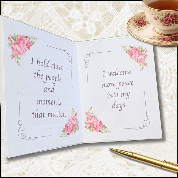 An open personalized greeting card with pink floral corner accents and printed quotes, styled on a cream lace surface with a teacup and gold pen nearby.