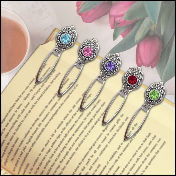 Five ornate silver metal bookmarks with colorful gemstone tops are arranged on an open book, with pink tulips and a cup of tea in the background.