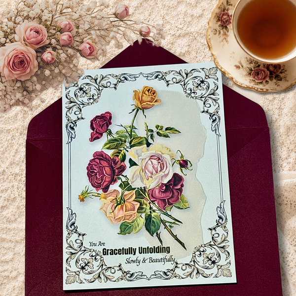 Vintage-inspired floral art card featuring roses and the words “Gracefully Unfolding, Slowly & Beautifully,” displayed on a burgundy envelope beside a teacup and lace background.