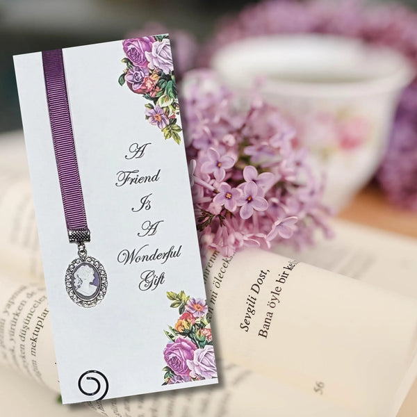 Victorian style purple ribbon bookmark with cameo charm on floral card reading “A Friend Is A Wonderful Gift,” displayed on an open book with lilac flowers and teacup in soft vintage setting