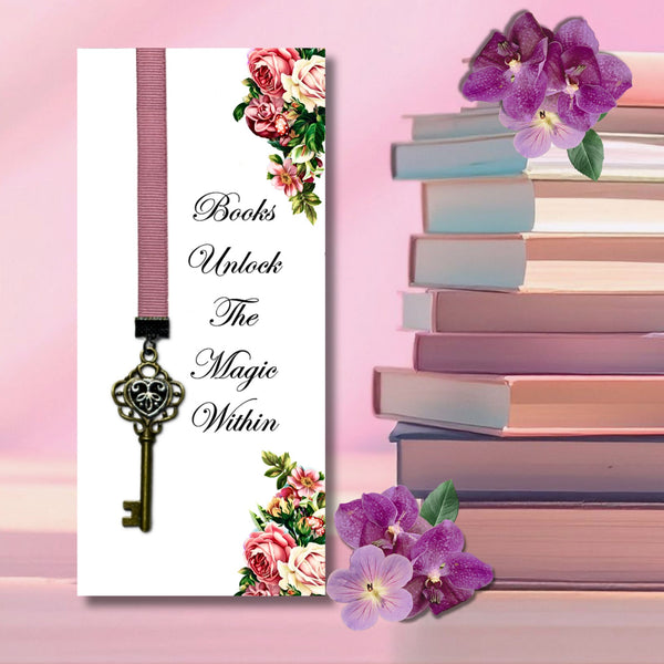 A white book bookmark card featuring a pink ribbon and antique gold key illustration, with the text 'Books Unlock The Magic Within' and floral corner accents. The card is styled on a pink background next to a stack of books and purple flowers.