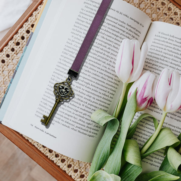 A purple fabric bookmark with a vintage-style metal key charm rests on an open book, styled with white and pink tulips on a woven mat.