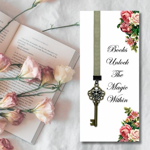 A decorative bookmark card displayed on a white fabric background next to an open book and pink flowers. The bookmark has a white background, a vertical olive green ribbon, an antique-style key hanging from the ribbon, and floral illustrations in the top right and bottom corners.