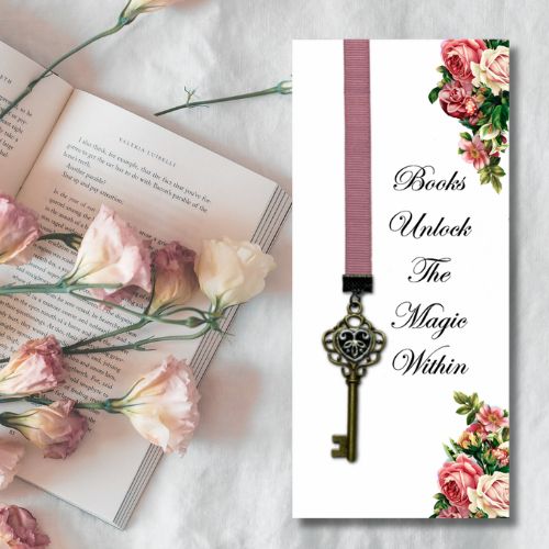 A decorative bookmark card displayed on a white fabric background alongside an open book and pink flowers. The bookmark has a white base, a vertical dusty pink ribbon, a vintage-style metal key hanging from the ribbon, and floral corner accents in pink and cream tones.