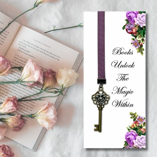 A personalized book bookmark with a purple ribbon, antique key charm, and floral corner accents, displayed next to an open book with pink roses on a white fabric background.