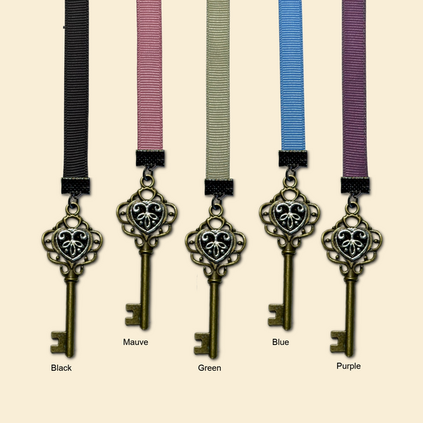Five decorative antique-style key charms with heart-shaped filigree tops are shown hanging from ribbed ribbon straps in different colors: black, mauve, green, blue, and purple. Each key is labeled beneath with its corresponding color name, and the background is a plain warm beige.