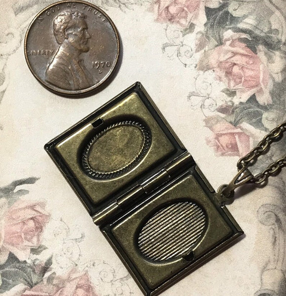 An antique-style bronze locket pendant shown open on a floral fabric background, with a U.S.