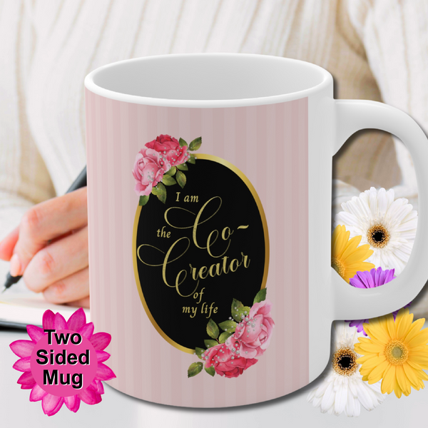 ceramic mug with saying" I am the co-creator of my life" written in gold lettering on a black oval with gold edge accented with large pink roses at the top and bottom of the oval on a background of pink stripes. Person writing in the background.