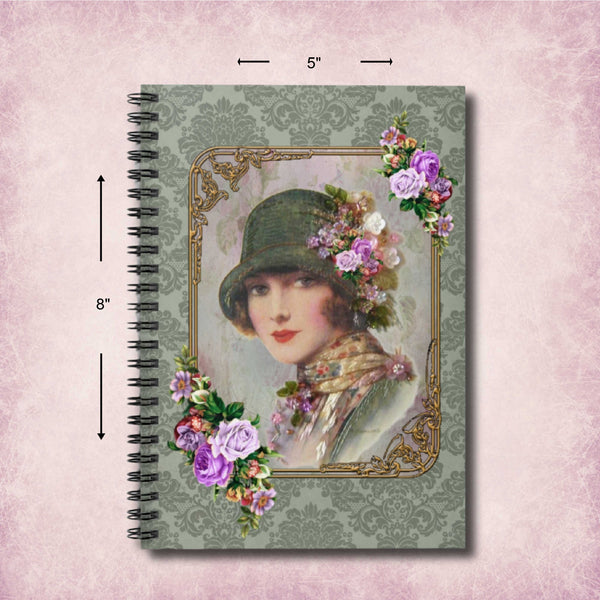 A spiral-bound notebook or journal shown front-facing on a pink textured background, with dimension arrows and labels indicating 8 inches tall, 5 inches wide, and the spiral spine. The cover features a vintage portrait of a woman in a green hat surrounded by purple and pink flowers, set within an ornate gold frame on a muted damask-patterned background.