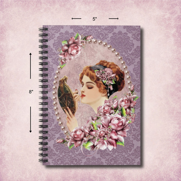 A spiral-bound notebook with a vintage-style cover illustration of a woman holding a mirror, surrounded by pink flowers and pearls on a lavender damask background. Black dimension arrows and labels indicate 8 inches tall by 5 inches wide on a pale pink textured backdrop.