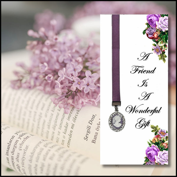A vertical white bookmark card with a purple ribbon and oval silver-toned pendant, shown beside an open book with pink flowers. The card reads, 'A Friend Is A Wonderful Gift' in black script, with purple and multicolor floral illustrations at the top and bottom corners.
