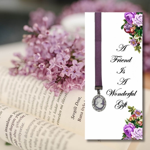 A memorial-style bookmark card with a purple ribbon and oval silver pendant, shown beside an open book with pink flowers. The card reads, 'A Friend Is A Wonderful Gift' with floral decorations in the corners.