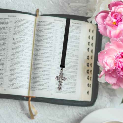 A silver-toned cross bookmark with a black ribbon lies on an open Bible, styled with pink flowers on a white textured surface.