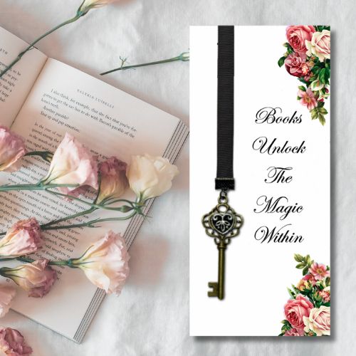 A decorative bookmark card displayed on a white fabric background next to an open book and pink flowers. The bookmark has a white background, black ribbon, a vintage key pendant, and floral corner accents with the text 'Books Unlock The Magic Within'.