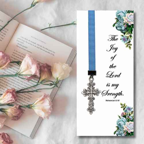 A vertical white prayer card or bookmark with a blue ribbon and silver cross, printed with the Bible verse 'The Joy of the Lord is my Strength' and floral corner designs, styled beside an open book with pink and cream flowers on a white fabric background.