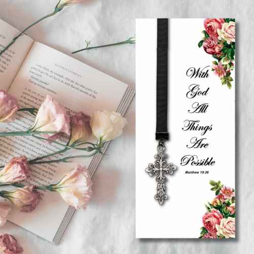 A vertical white prayer card with a black ribbon and silver cross pendant, printed with the phrase 'With God All Things Are Possible Matthew 19:26' and decorated with pink and green floral illustrations in the corners. The card is shown beside an open book with pink flowers on a light fabric background.