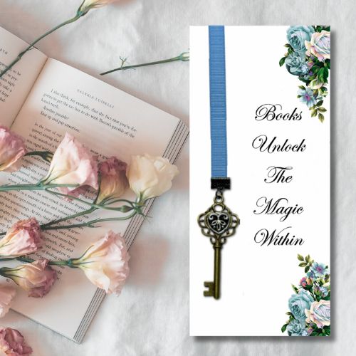 A decorative bookmark or card featuring a blue ribbon loop with an antique-style key charm, shown beside an open book and pink flowers on a light fabric background. The card reads, 'Books Unlock The Magic Within' with floral accents in the corners.