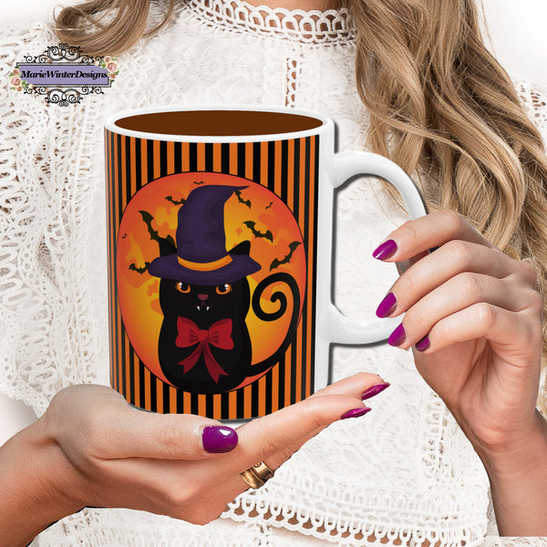 Hand with dark rose nail polish holding Halloween mug with black cat wearing a purple pointed had and a red bow around neck in side a circle with bats flying behind on an orange and black stripe background with a white handle