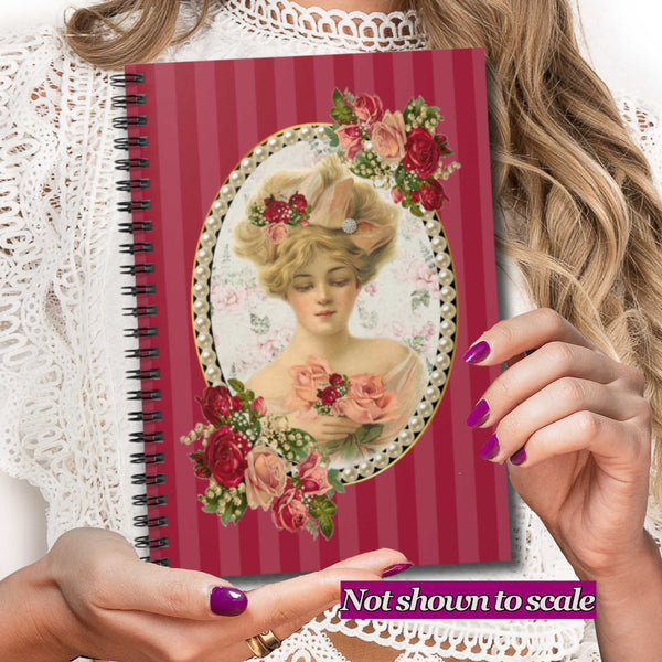 A person holds a spiral-bound notebook with a vintage-style illustration of a woman in an ornate floral frame. The notebook cover has red vertical stripes and pink floral accents.