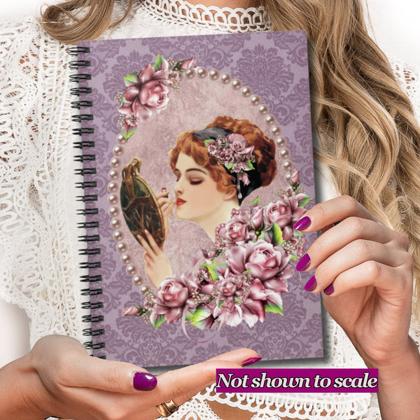 A person holds a spiral-bound notebook with a vintage-style illustration of a woman in profile, surrounded by pink flowers and a pearl-like border on a lavender damask background. The person wears a white lace top, and a purple 'Not shown to scale' banner appears at the bottom.
