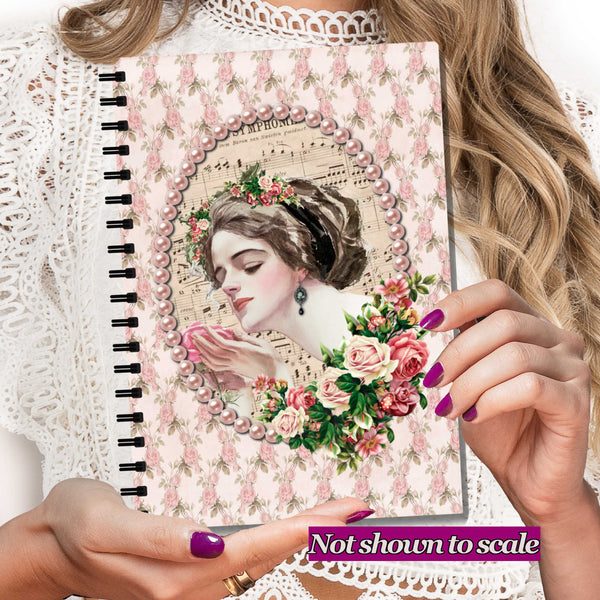A spiral-bound notebook with a vintage-style floral cover design featuring a woman in profile, pink roses, and sheet music, held by hands with purple nail polish. The person is wearing a white lace top.