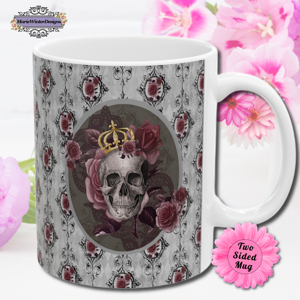 Ceramic mug with a skull wearing a gold crown accented with burgundy roses inside an oval with a light gray border, on a background of gray with black scrolls and burgundy roses with white handle. Pink flowers are behind the mug.